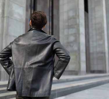 a mature man in a leather jacket is standing looking at the buildings in the city, the view from the back