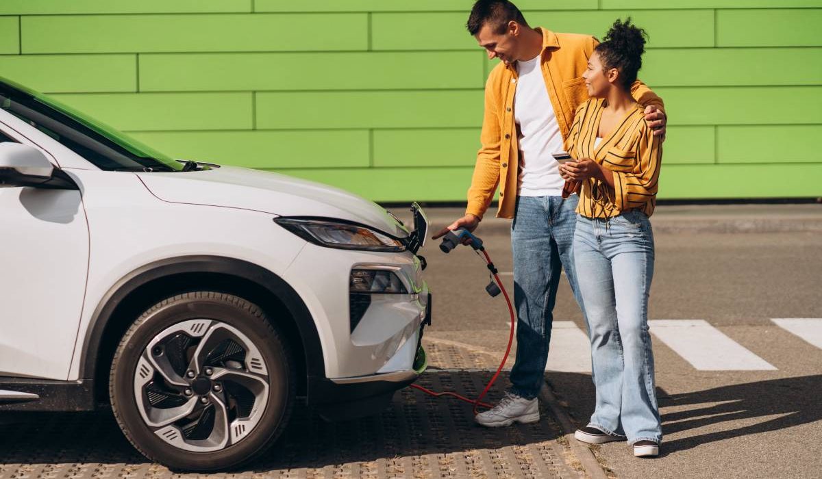 Young couple charging electric vehicle at EV charging station, using mobile phone