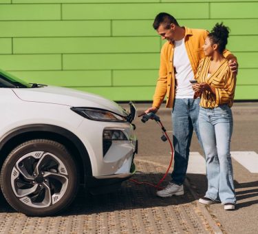 Young couple charging electric vehicle at EV charging station, using mobile phone