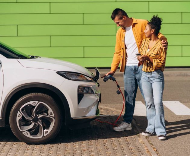 Young couple charging electric vehicle at EV charging station, using mobile phone